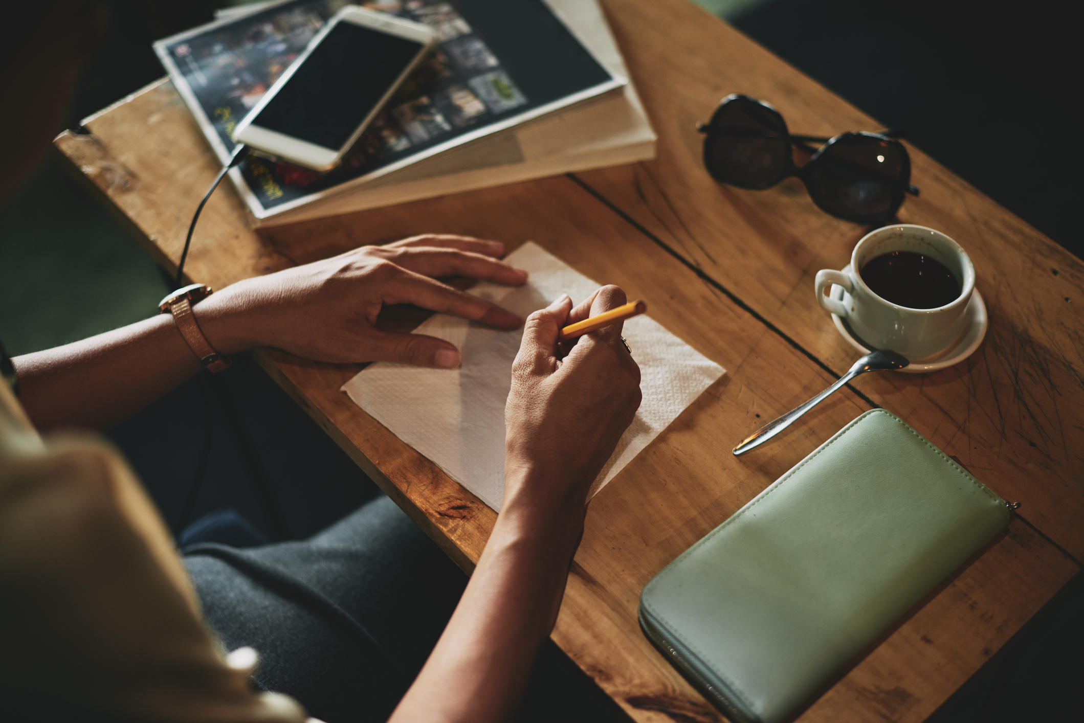 A person sits at a wooden desk next to a tea cup and a cellphone while writing on a napkin with a pencil