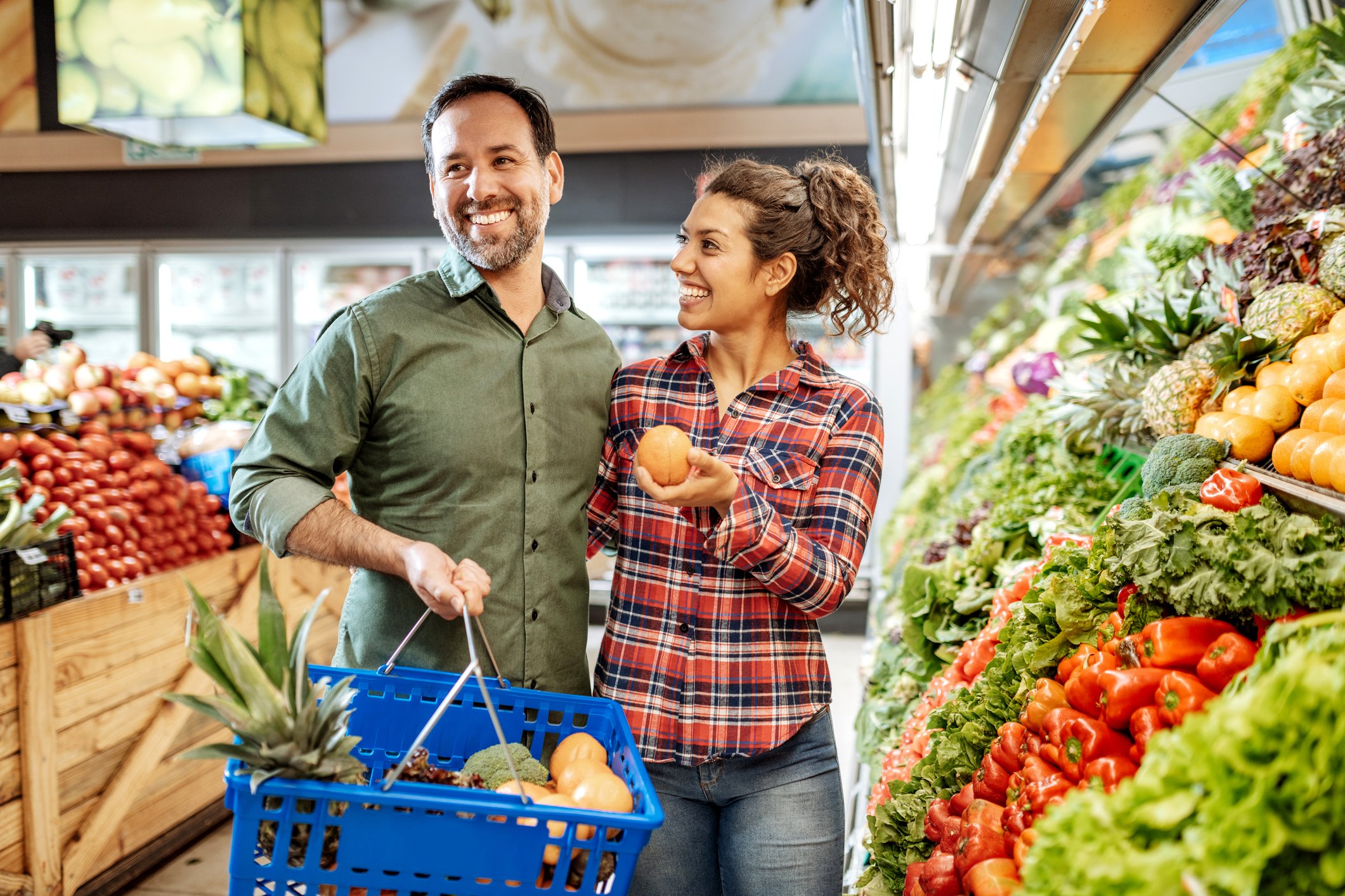 A man and woman smile in a grocery store while loading produce into a handheld cart