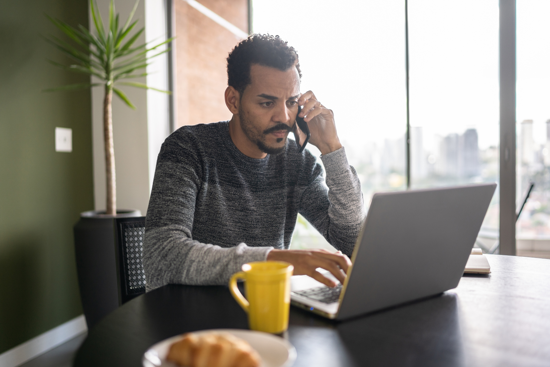 A man sits at a table and talks on a mobile phone while looking at a laptop