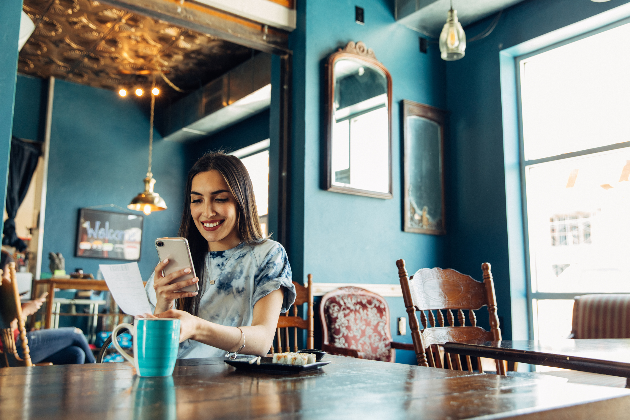 A woman sits in a restaurant and smiles while taking a photo of the bill with her phone